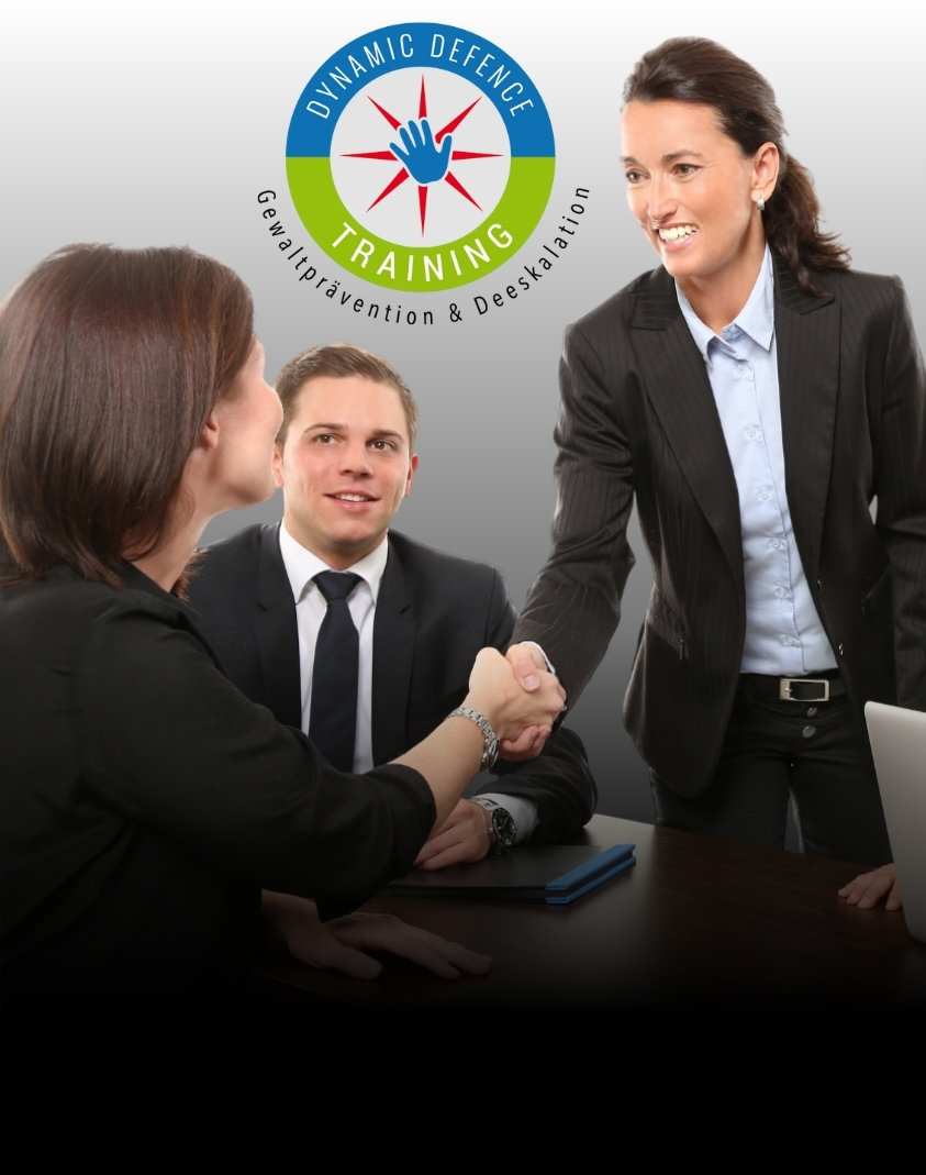 2 professionell gekleidete Damen schütteln sich die Hand. Ein Mann lächelt in die Kamera